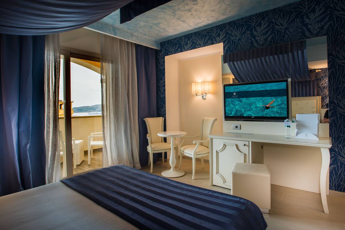 A hotel room with a bed, a desk with a computer, a chair, a small table, and a view of the sea.