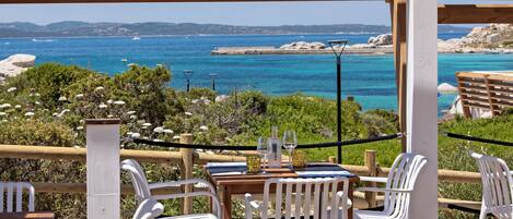 Un espacio al aire libre con sillas de plástico blanco y una mesa de madera, con vistas a la costa, aguas azules cristalinas y formaciones rocosas a lo lejos.
