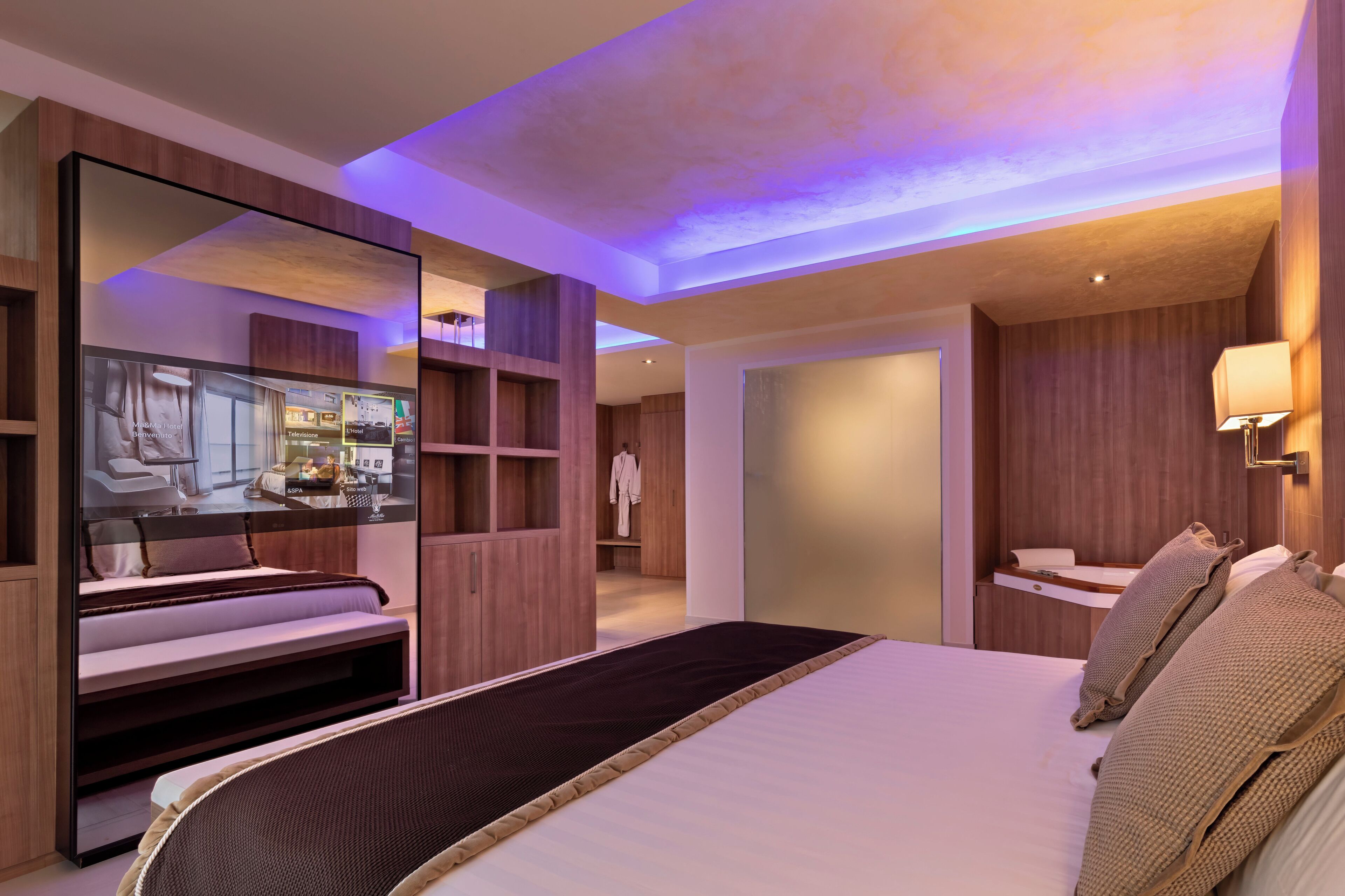A modern hotel room with a large bed, a mirror, and a television displaying the hotel's amenities.