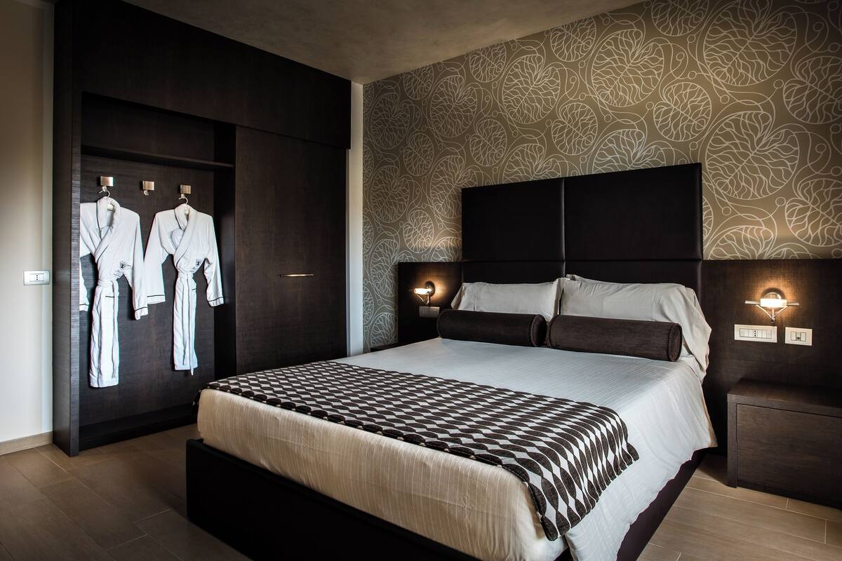 A modern hotel room with a large bed, dark headboard, patterned wallpaper, and two bedside lamps.