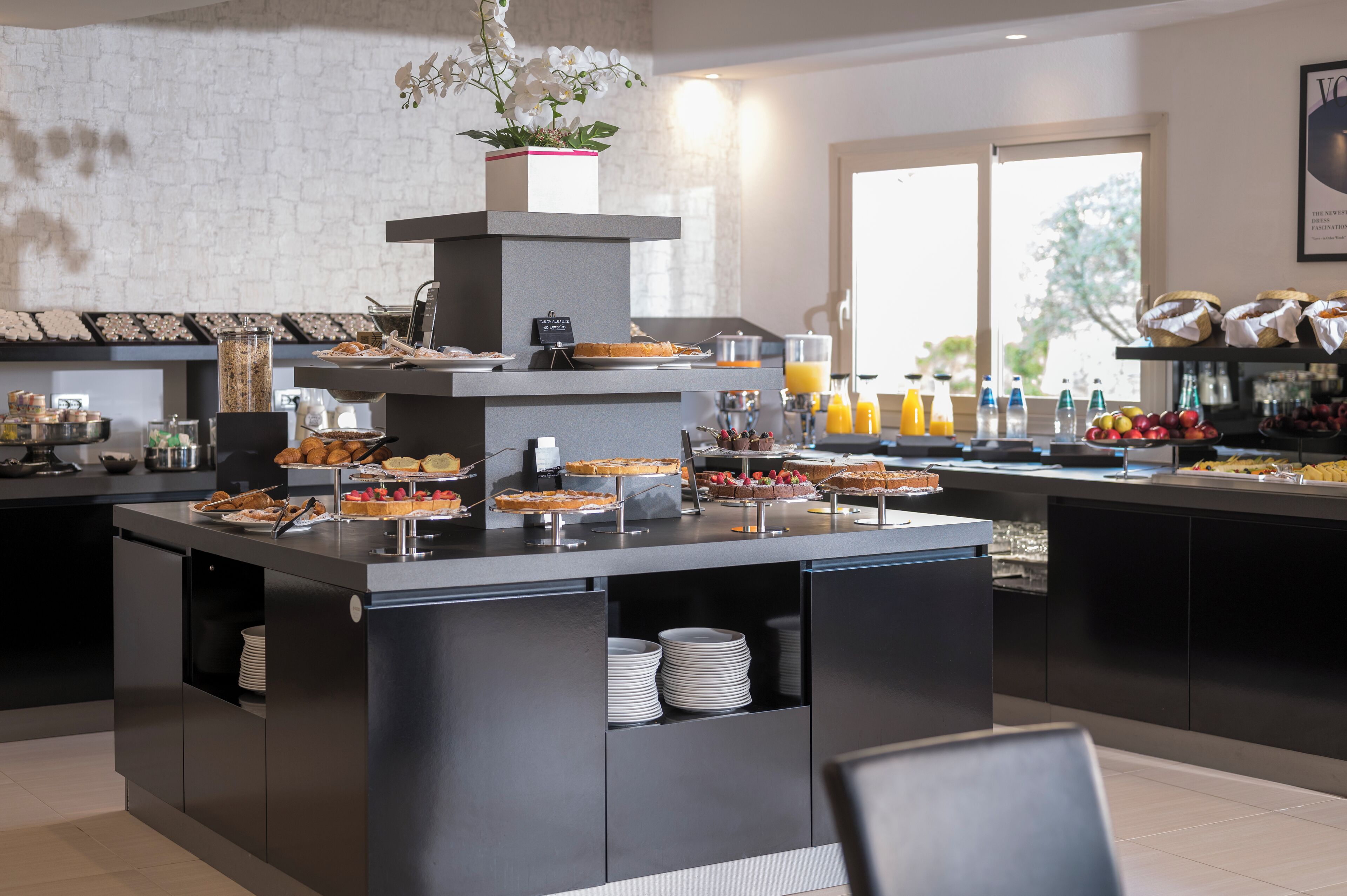 A modern breakfast bar with a variety of pastries and beverages, a coffee station, and a view of the outdoors through large windows.