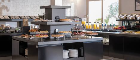 A modern breakfast bar with a variety of pastries and beverages, a coffee station, and a view of the outdoors through large windows.