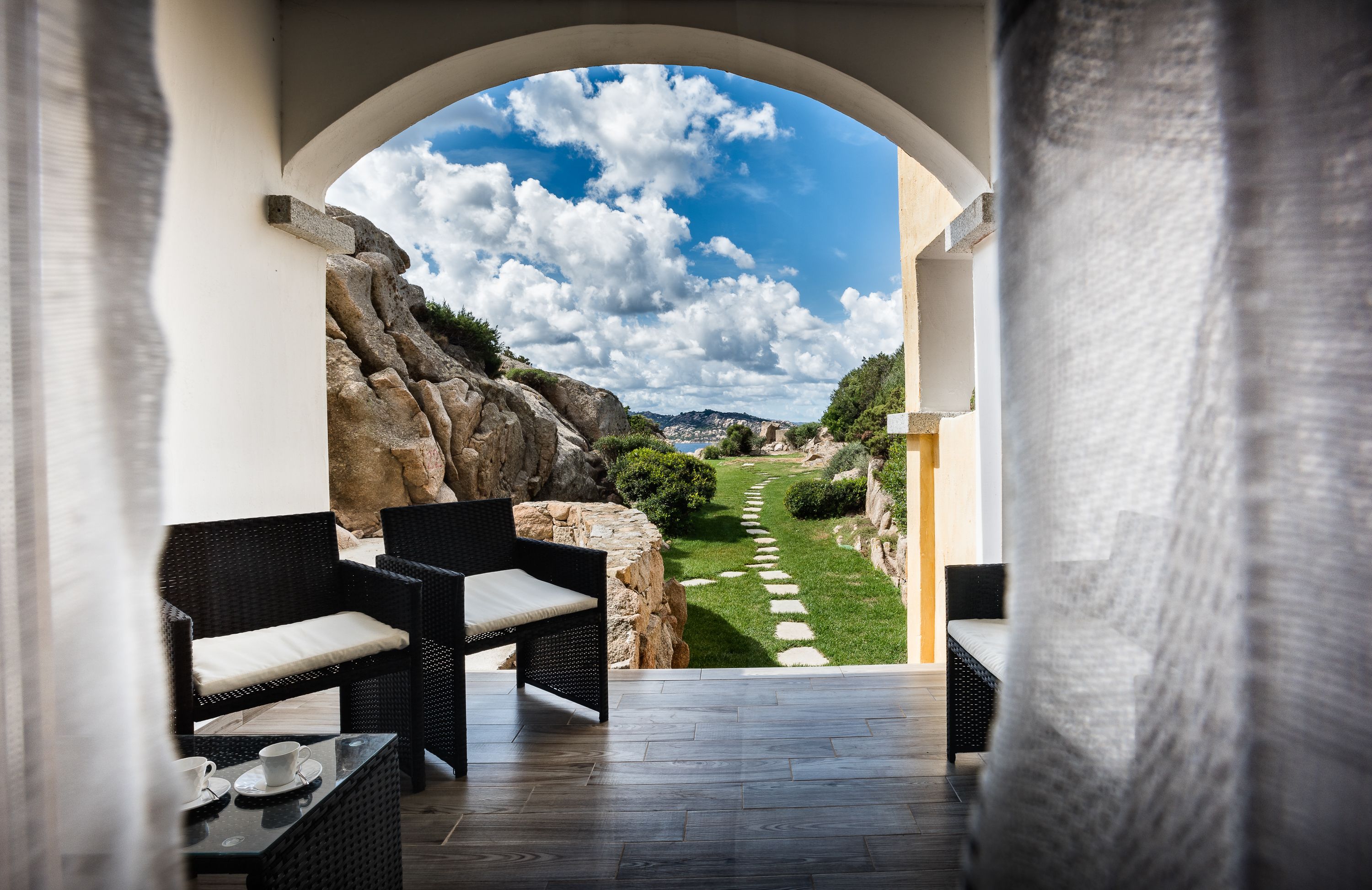 A covered outdoor seating area with wicker furniture and a view of a landscaped garden.