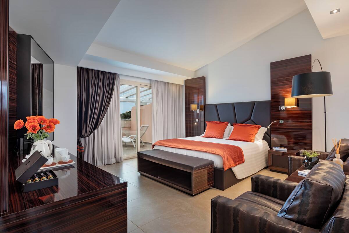 A modern hotel room with a large bed, a desk with a mirror, a sofa, and a dining area with a table and chairs.