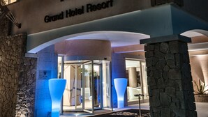 Exterior do Grand Hotel Resort, com uma placa iluminada em destaque, fachada de pedra e entrada moderna com portas de vidro.