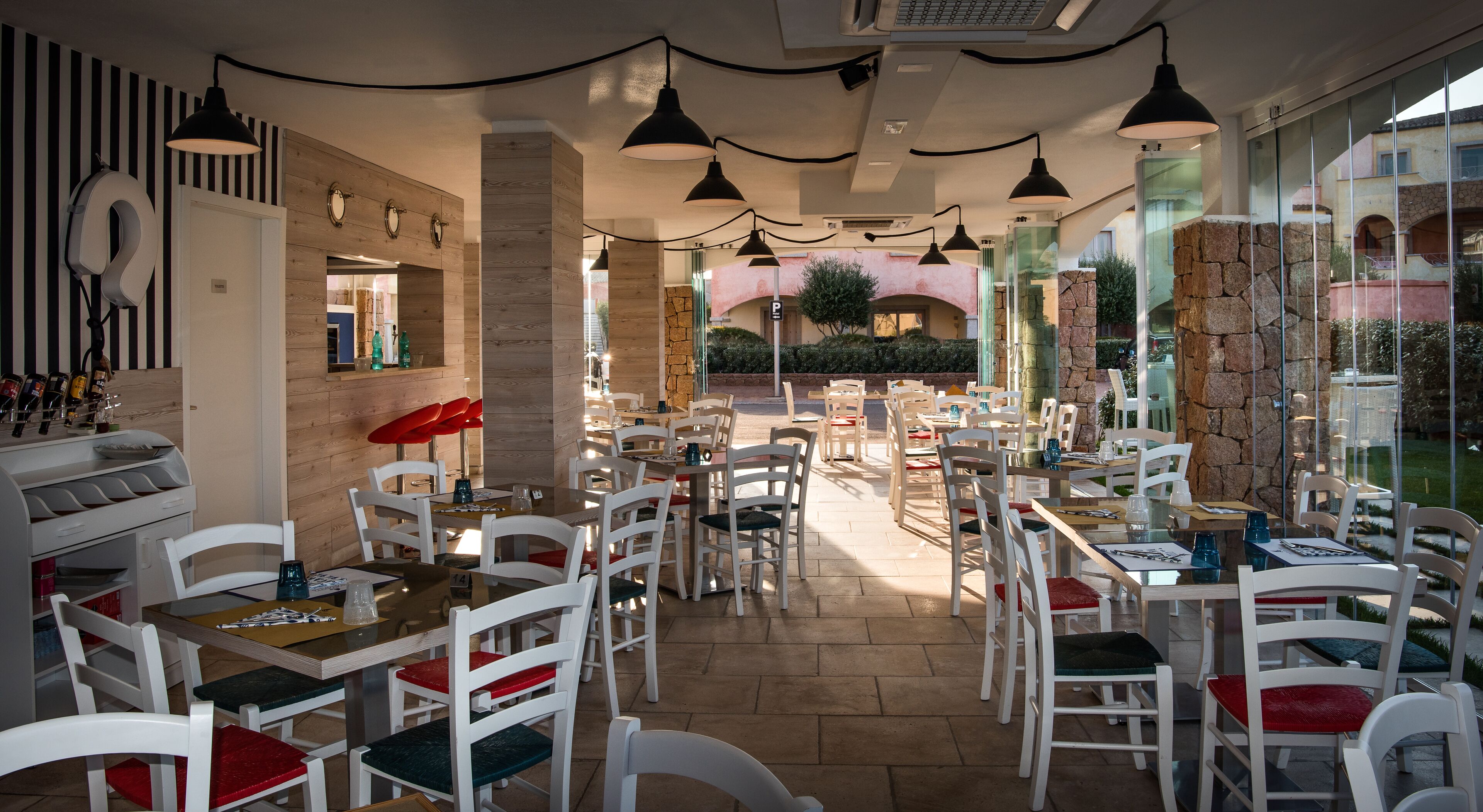 Un restaurant avec des chaises et des tables blanches, ainsi que des suspensions noires. On y trouve un mur en pierre et une porte vitrée donnant sur l’extérieur.