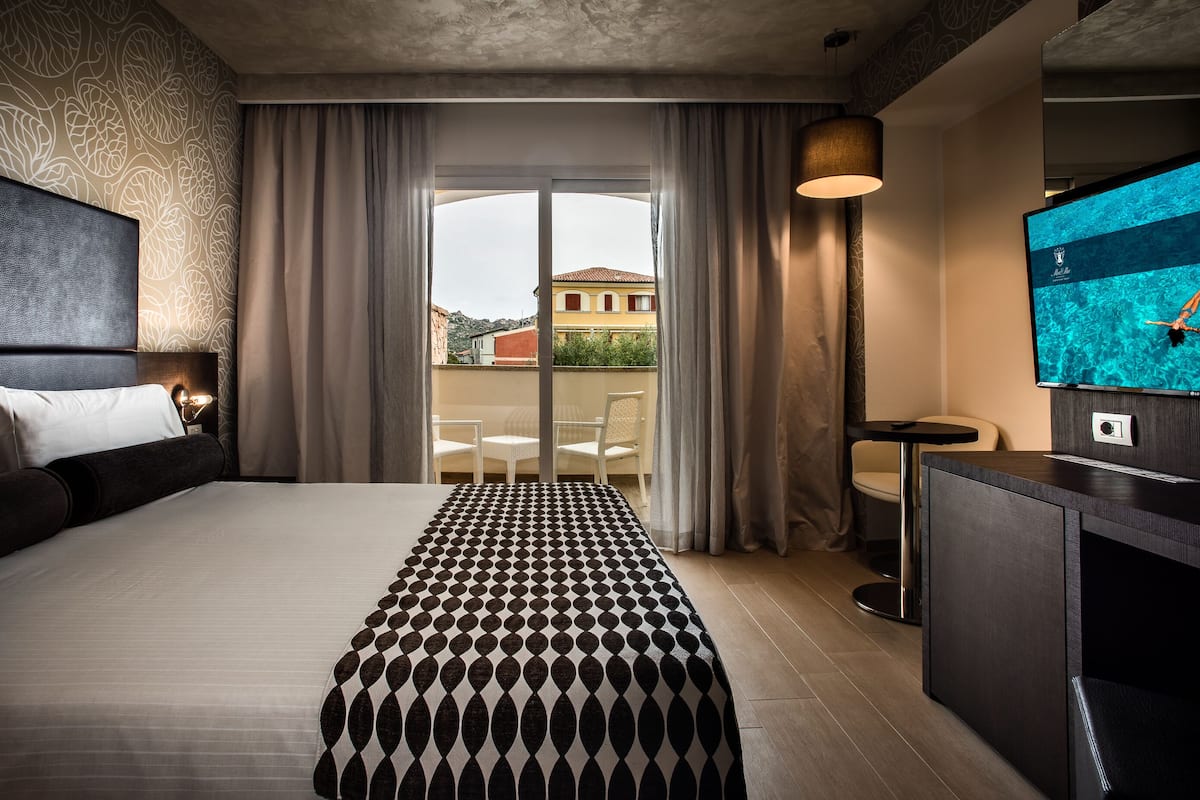 A hotel room with a large bed, a small table, a TV, and a balcony with curtains.