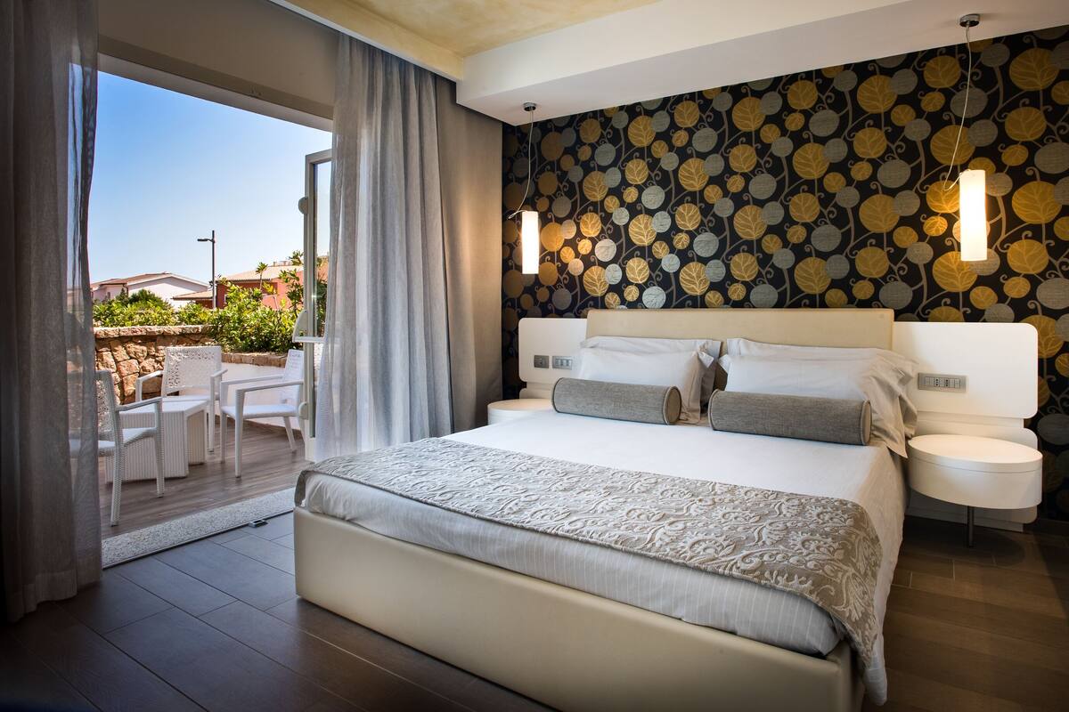 A modern hotel room with a large bed, a balcony with outdoor seating, and a decorative leaf-patterned wall.