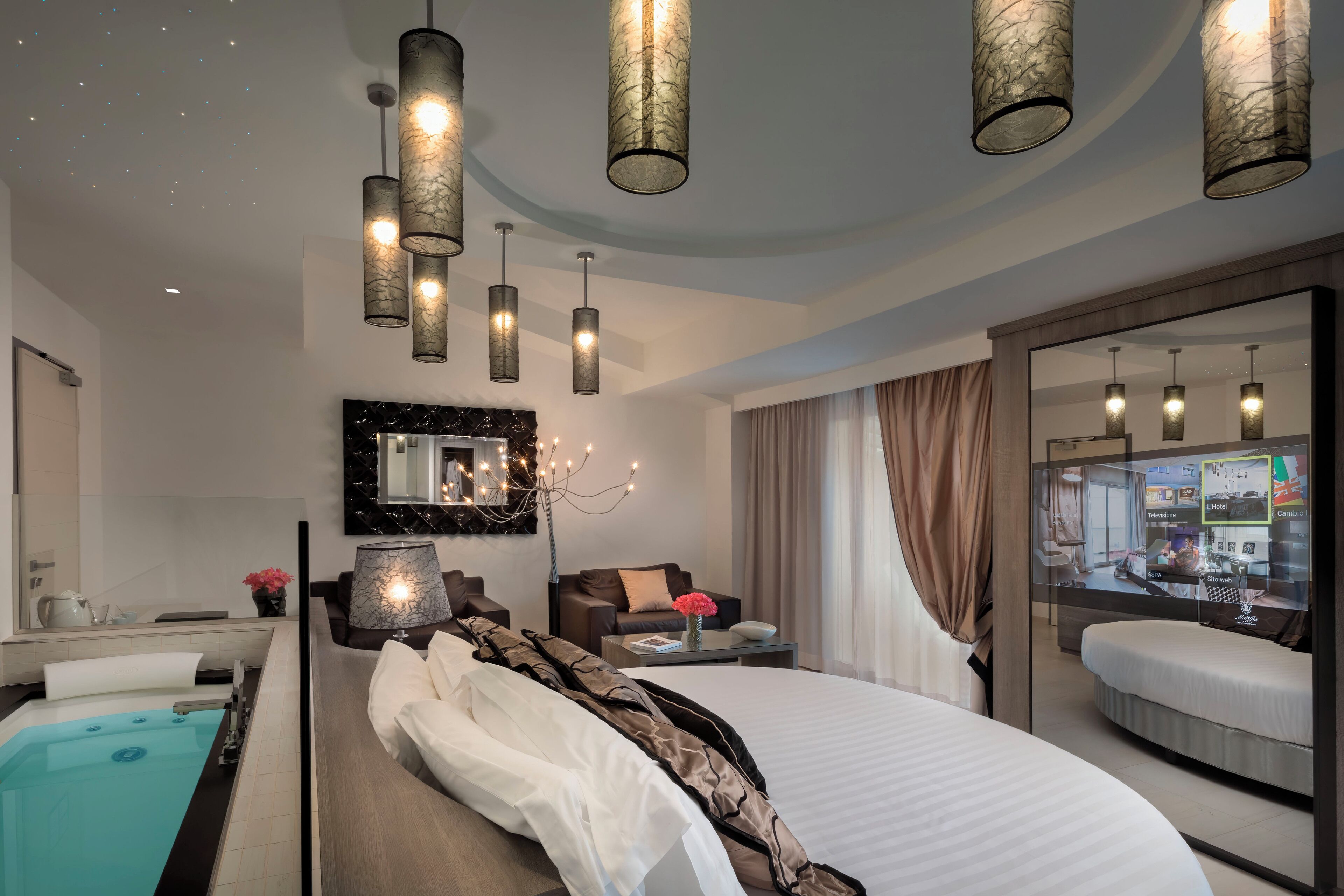 A modern hotel room with a large bed, a separate sitting area with a sofa, a small table, a large mirror, and a view of the city through a glass wall.