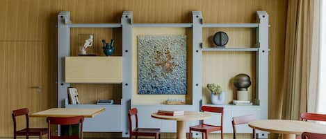 A modern dining area with wooden tables and red chairs, a large abstract painting on the wall, and a bookshelf with decorative items.