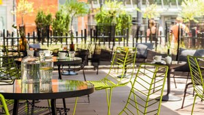 An outdoor cafe with round tables and green chairs, surrounded by a glass fence and greenery.