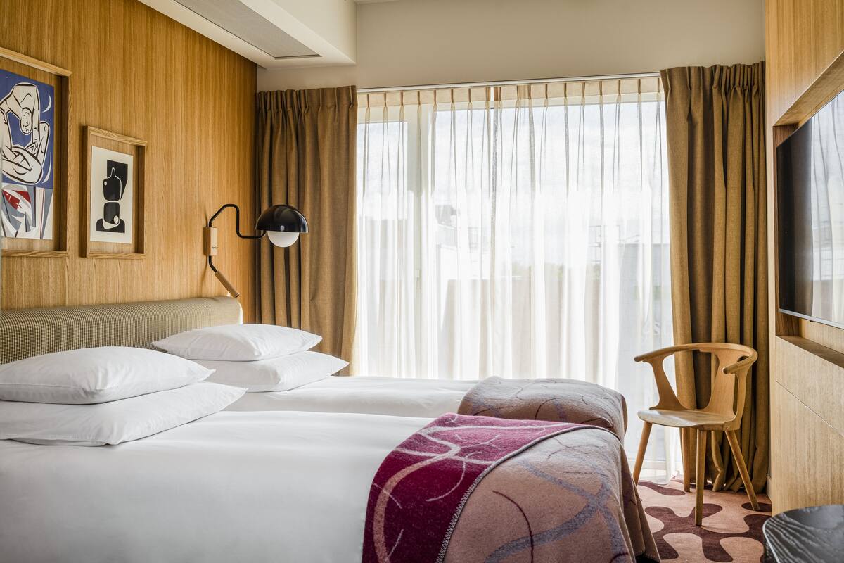 Een hotelkamer met twee bedden, een houten hoofdbord, een televisie, een stoel en een groot raam met gordijnen.