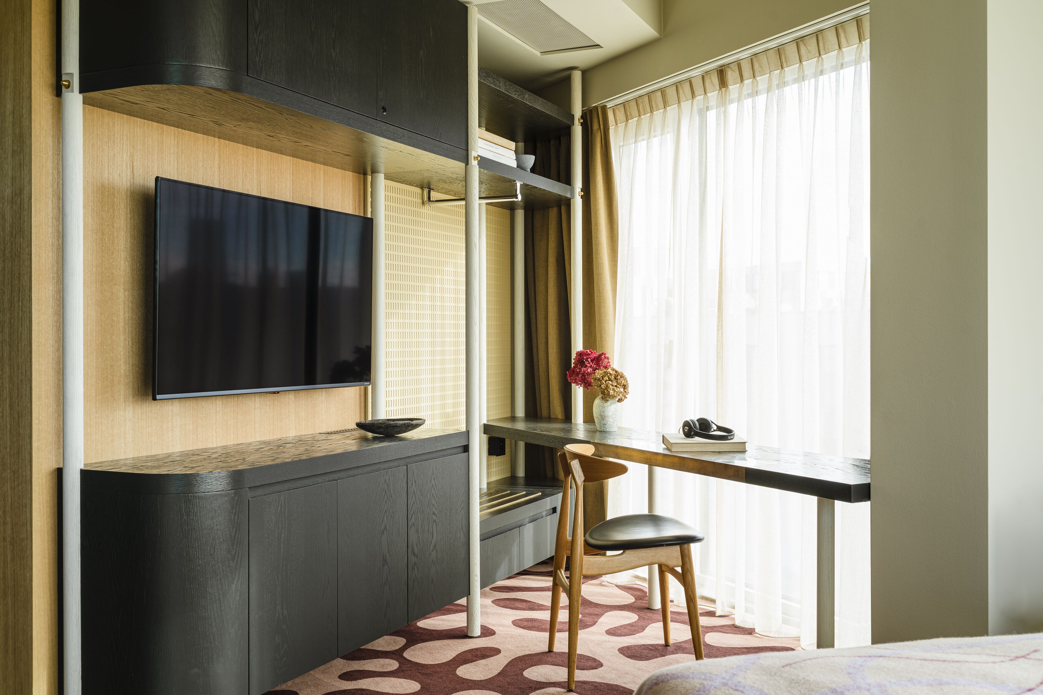 A compact hotel room with a TV mounted on the wall, a small desk with a chair, and a window with curtains.