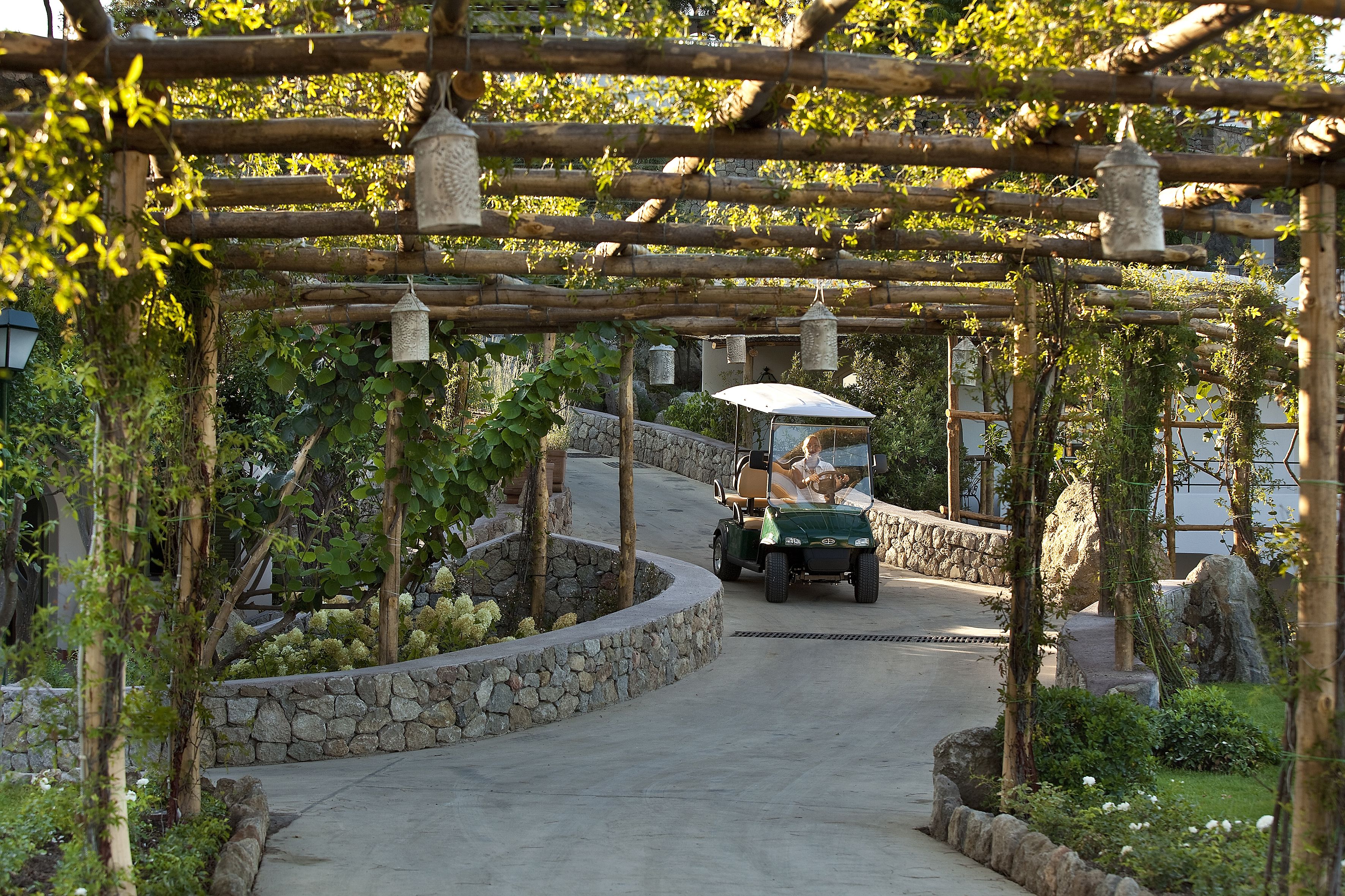 A pathway with a stone wall on one side and a wooden pergola with hanging lanterns.
