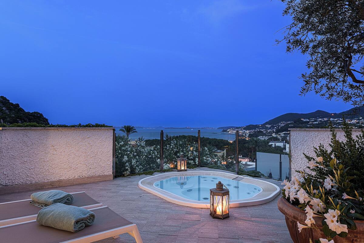 A rooftop pool area with a hot tub, lounge chairs, and a view of the sea and cityscape.