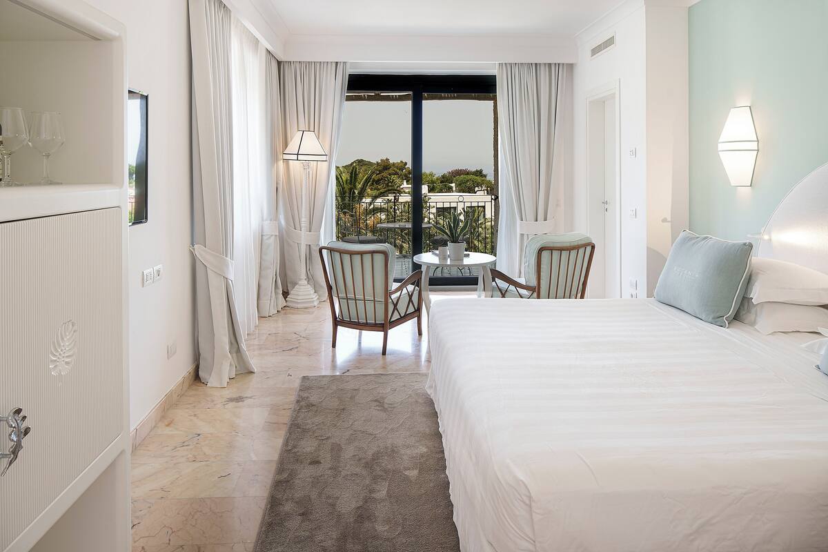 A hotel room with a bed, two chairs, a small table, a view of the outdoors, and a balcony with a railing.