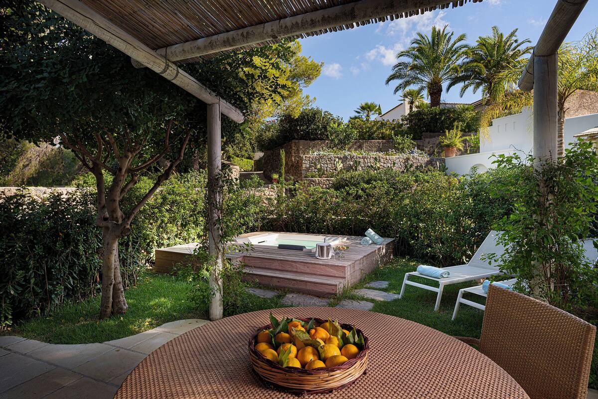 A patio with a woven table and chairs, a wicker basket of fruit, and a hot tub in the background.