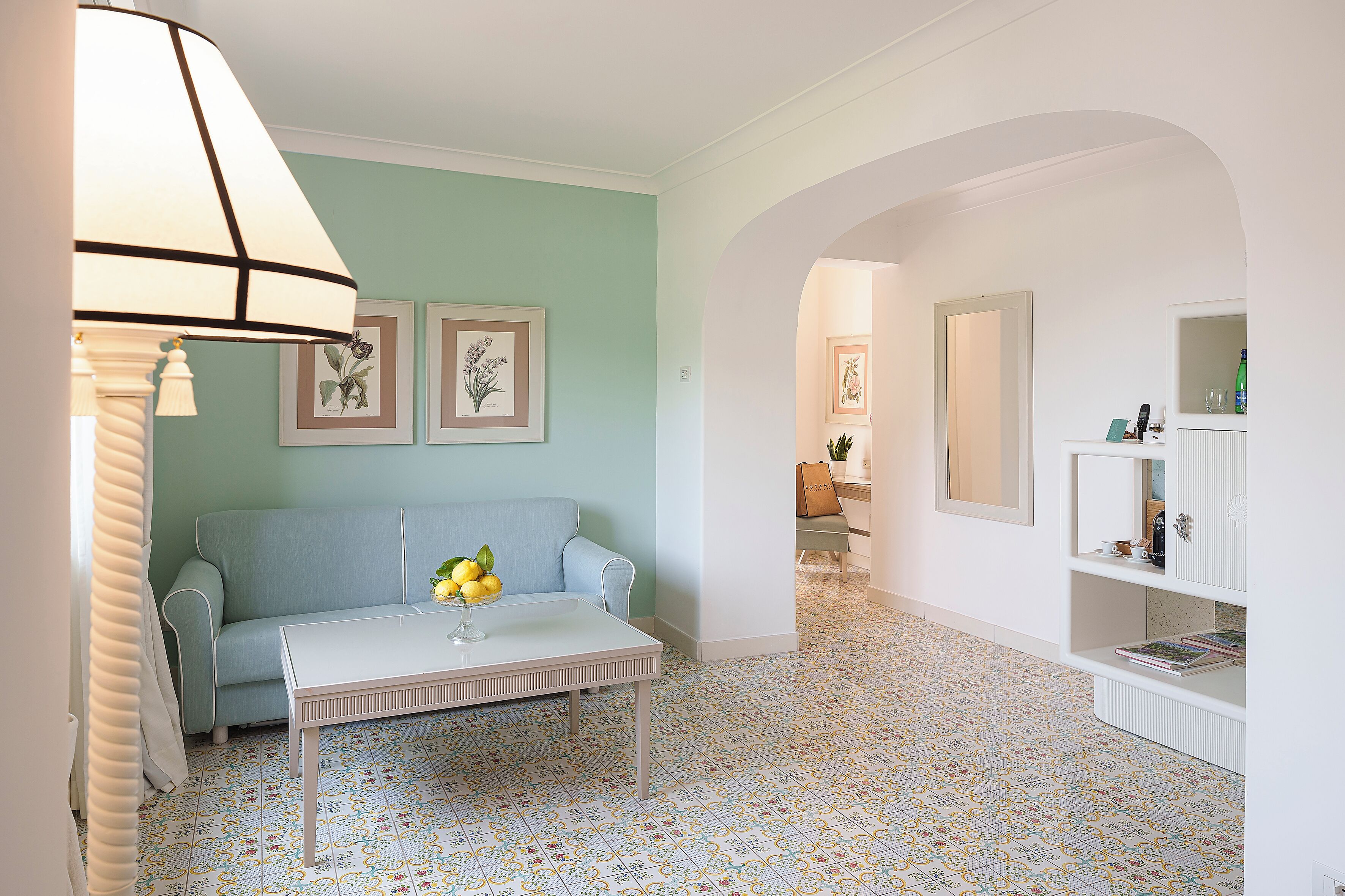 Una sala de estar con un sofá, una mesa de centro y una lámpara. En la pared hay cuadros enmarcados y una estantería con libros y objetos decorativos.