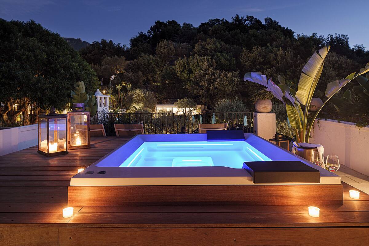 A heated outdoor pool area with wooden decking, surrounded by lounge chairs and candles, set against a backdrop of trees and a hill.