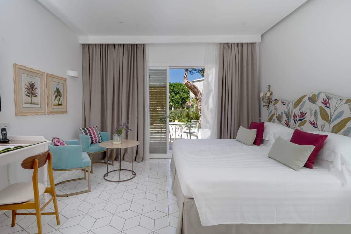 A modern hotel room with a bed, a desk, a chair, a small table, and a balcony with a view of greenery.