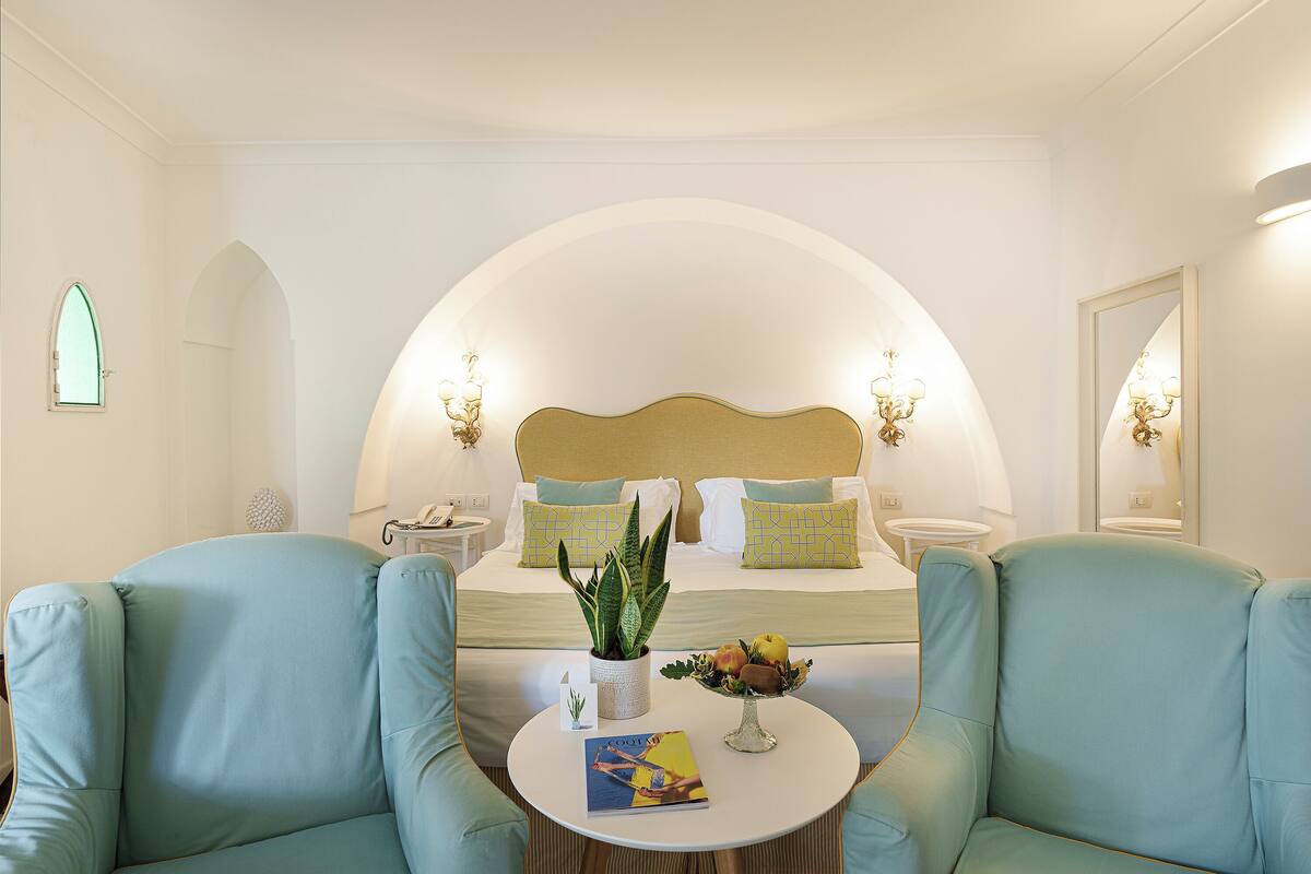 A hotel room with a bed, two armchairs, a round table with a magazine, a plant, and a fruit bowl.