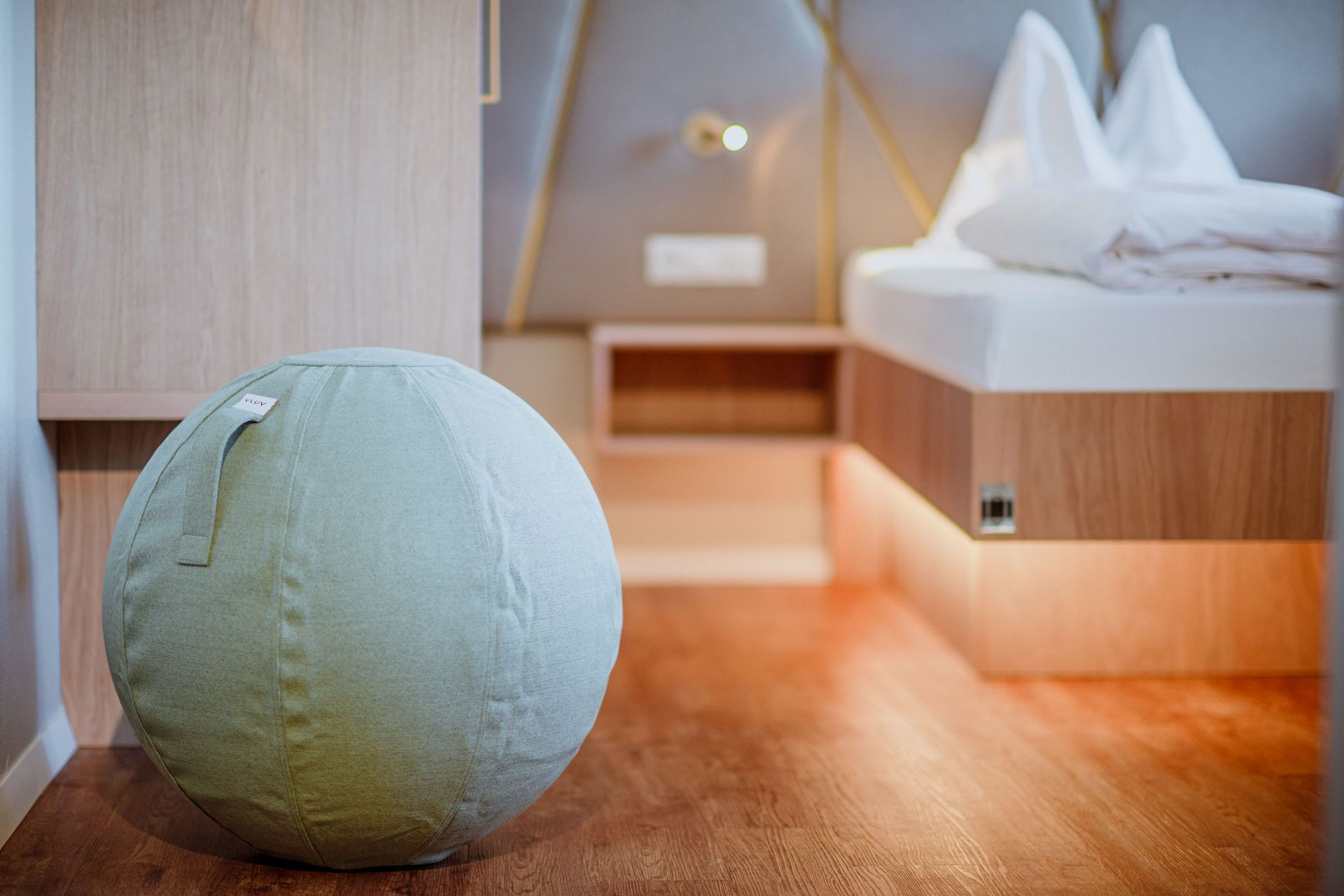A room with a round, light-colored bean bag chair, a wooden floor, a bed with white bedding, and a wooden wardrobe.