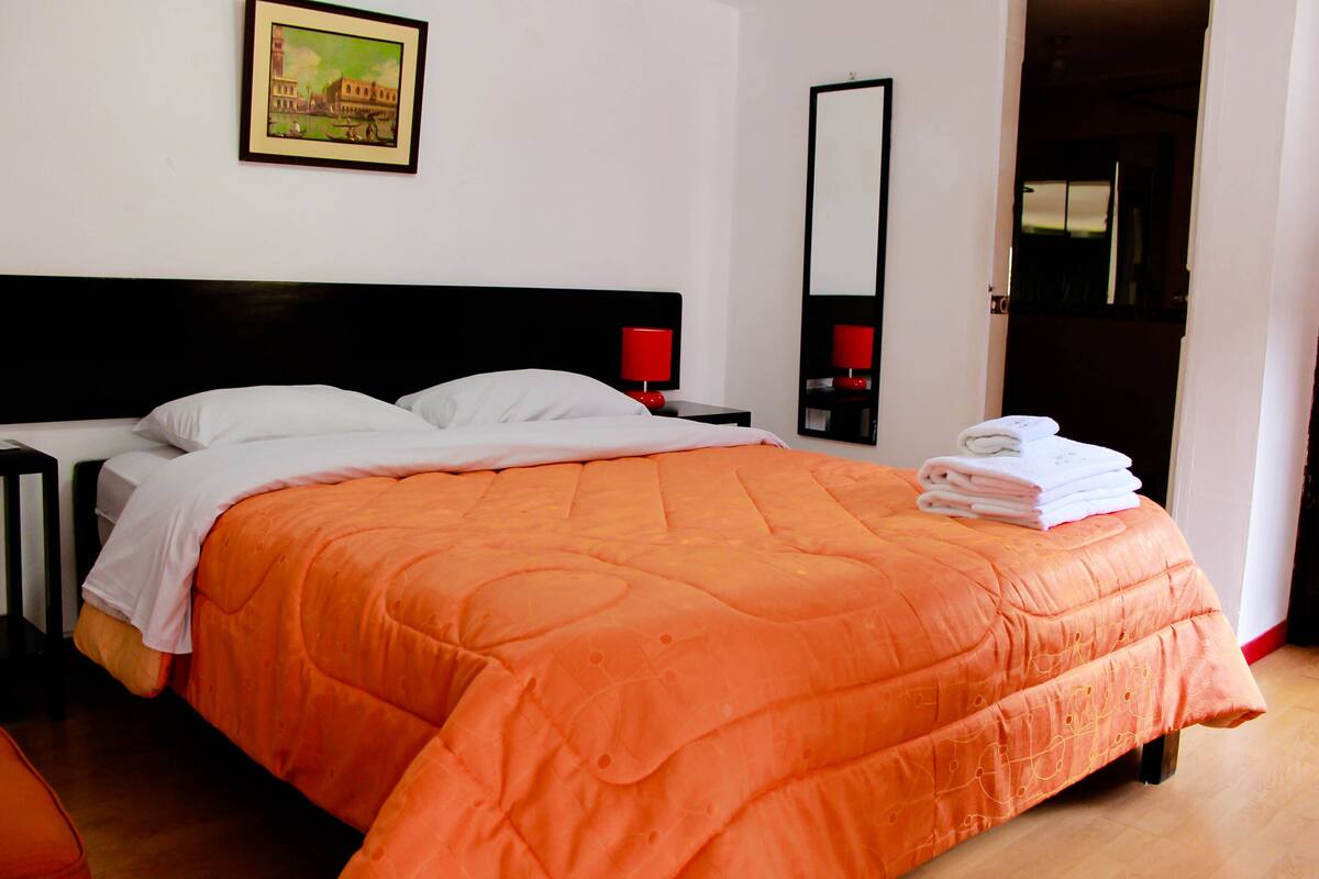 Una cama bien hecha con una colcha naranja, una almohada blanca y una lámpara roja sobre una mesita de noche.