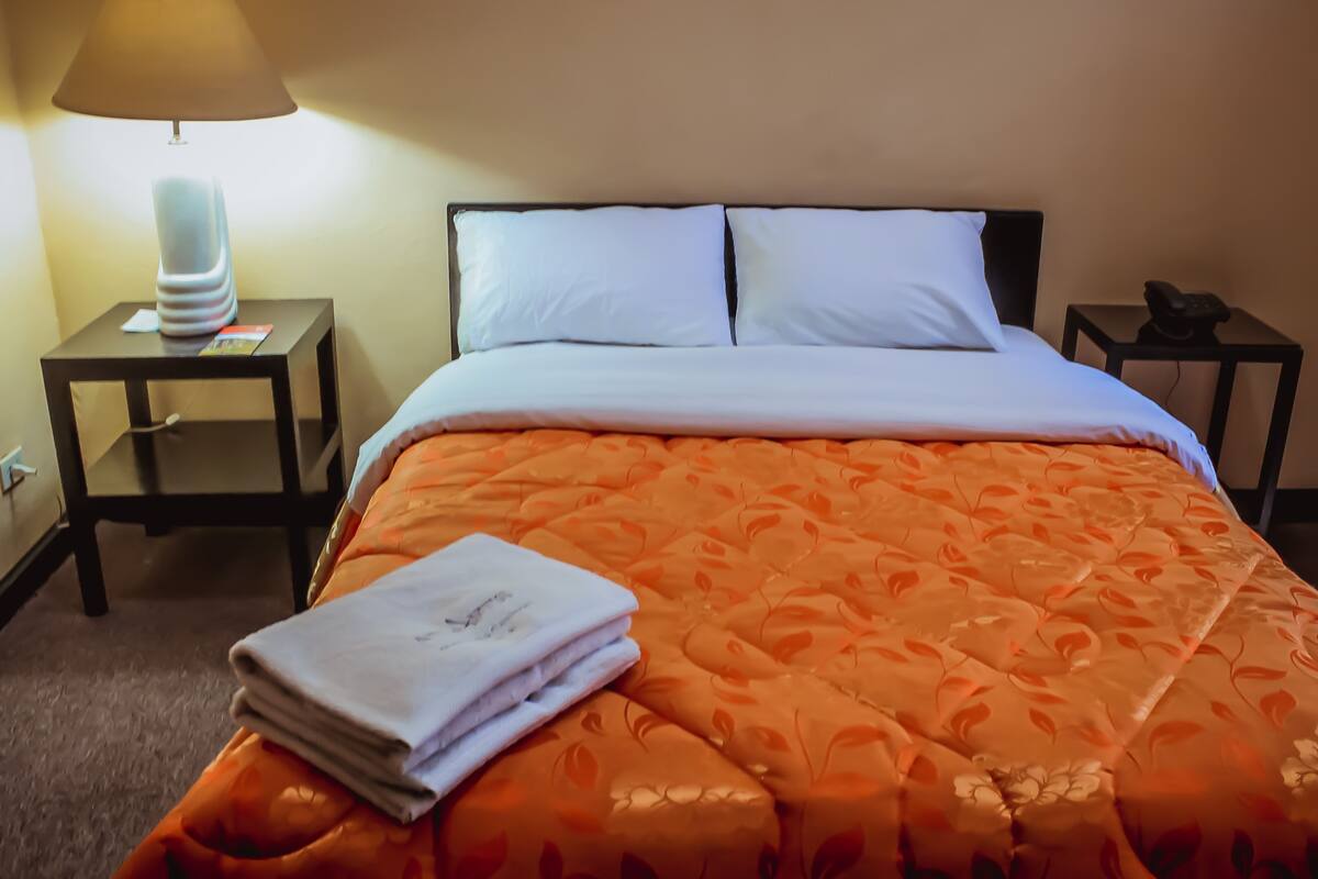 Una habitación de hotel con una cama, dos mesitas de noche, una lámpara y una toalla doblada.