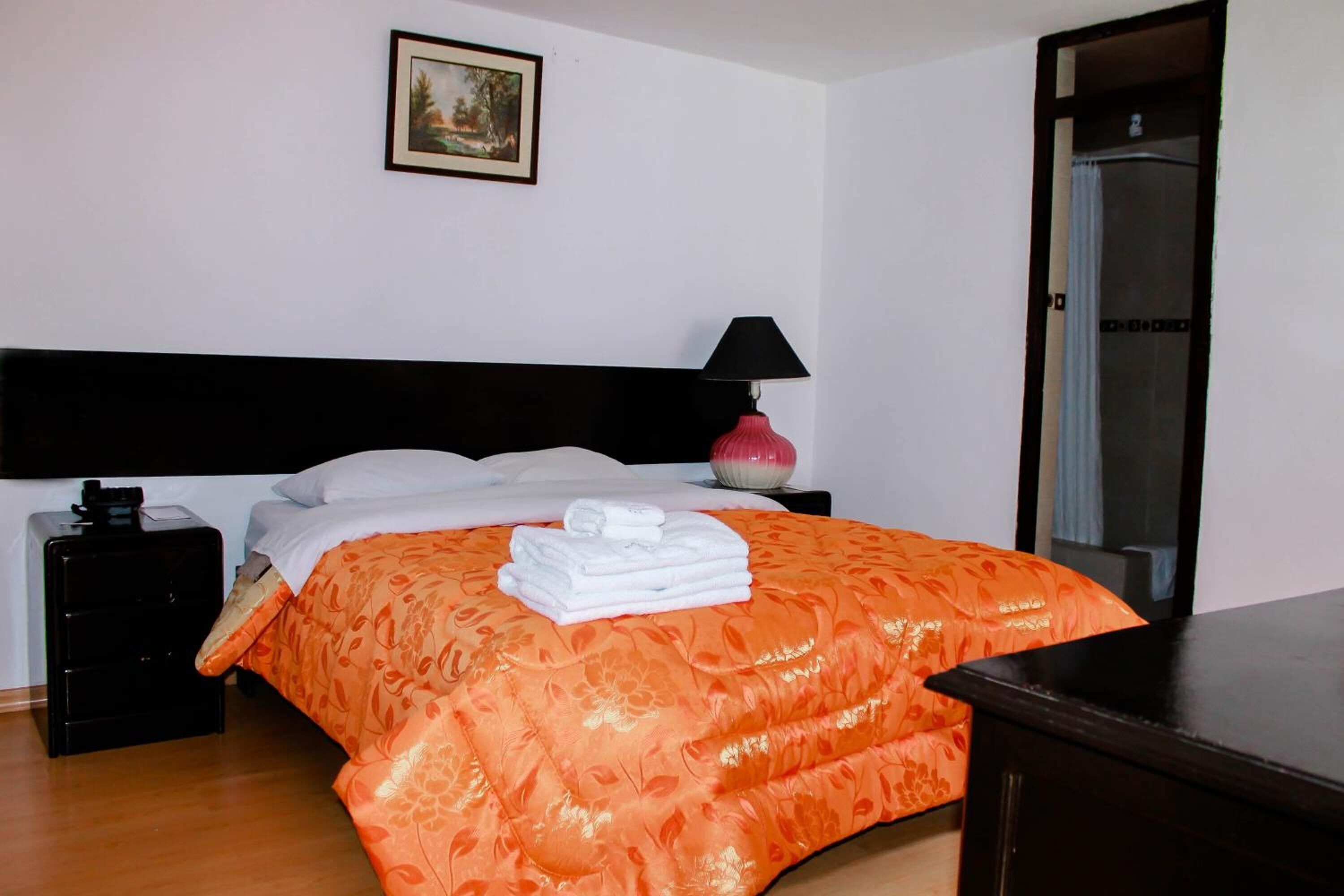 Una cama bien hecha con colcha naranja estampada, cabecera negra, lámpara de noche y un cuadro en la pared.