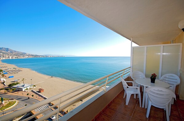 Un balcón con vistas a una playa y al mar. Hay sillas de plástico blanco y una mesa con un jarrón de flores.