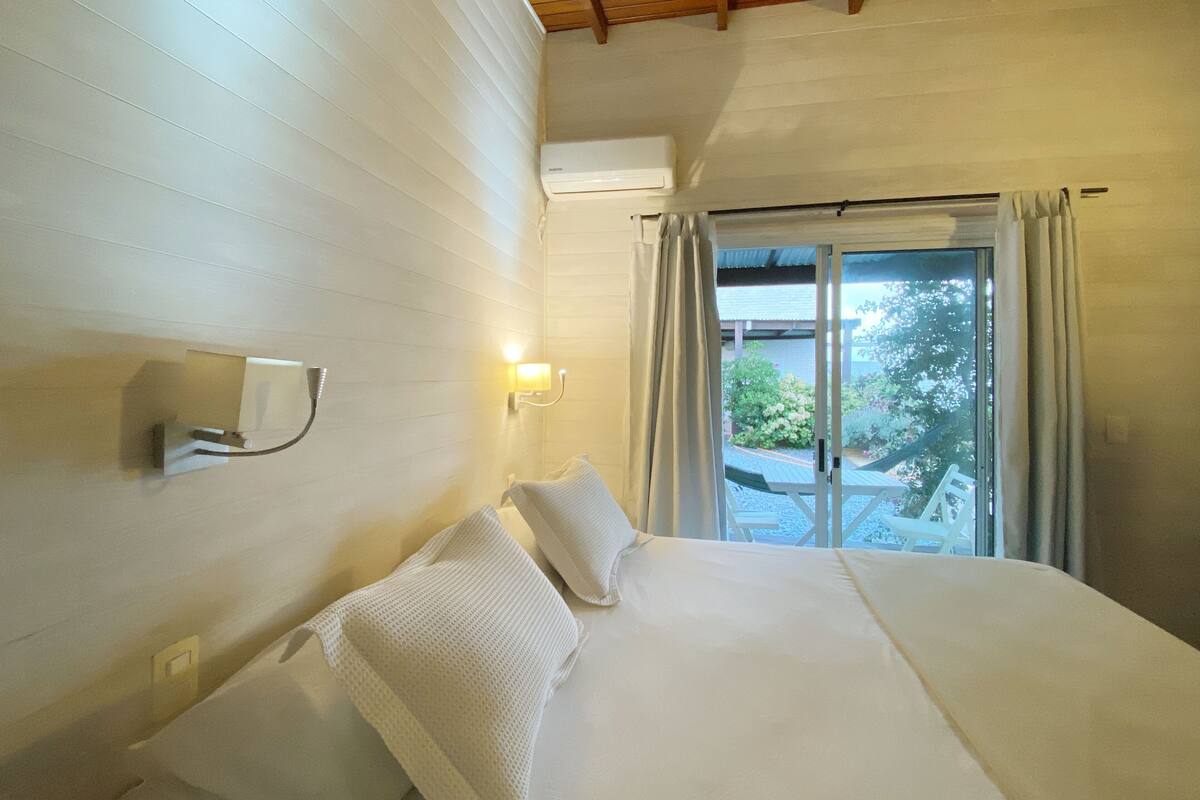 A hotel room with a large bed, a wall-mounted air conditioner, a window with curtains, and a view of the outdoors.