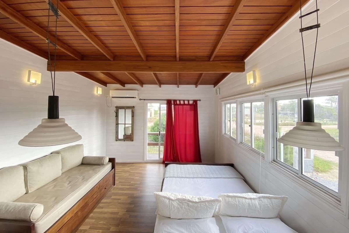 A bedroom with a wooden ceiling, a bed with white bedding, a sofa with cushions, and a window with a view of greenery.