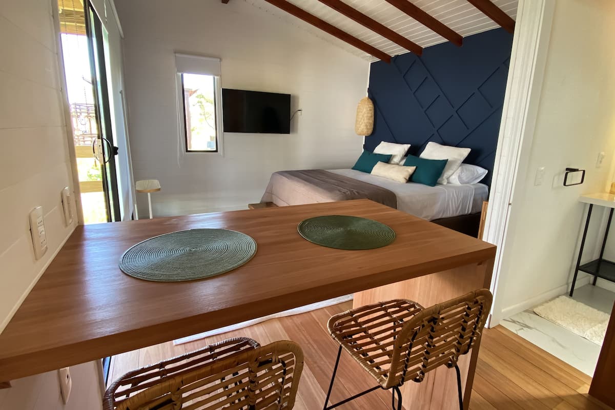 A modern room with a wooden dining table, two wicker chairs, a bed with blue and white bedding, and a TV mounted on the wall.