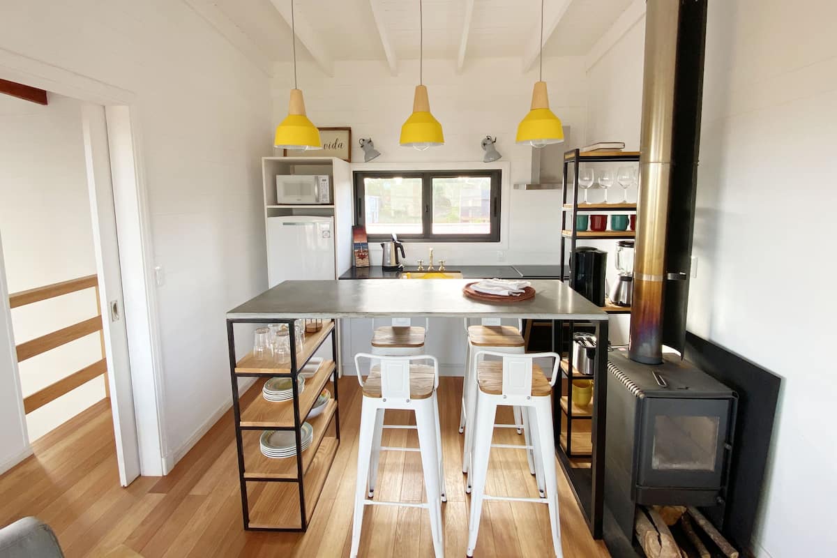 A modern kitchen with a central island, white bar stools, a wood-burning stove, and yellow pendant lights.