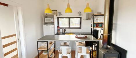 Et moderne køkken med en central ø, hvide barstole, en brændeovn og gule hængelamper.