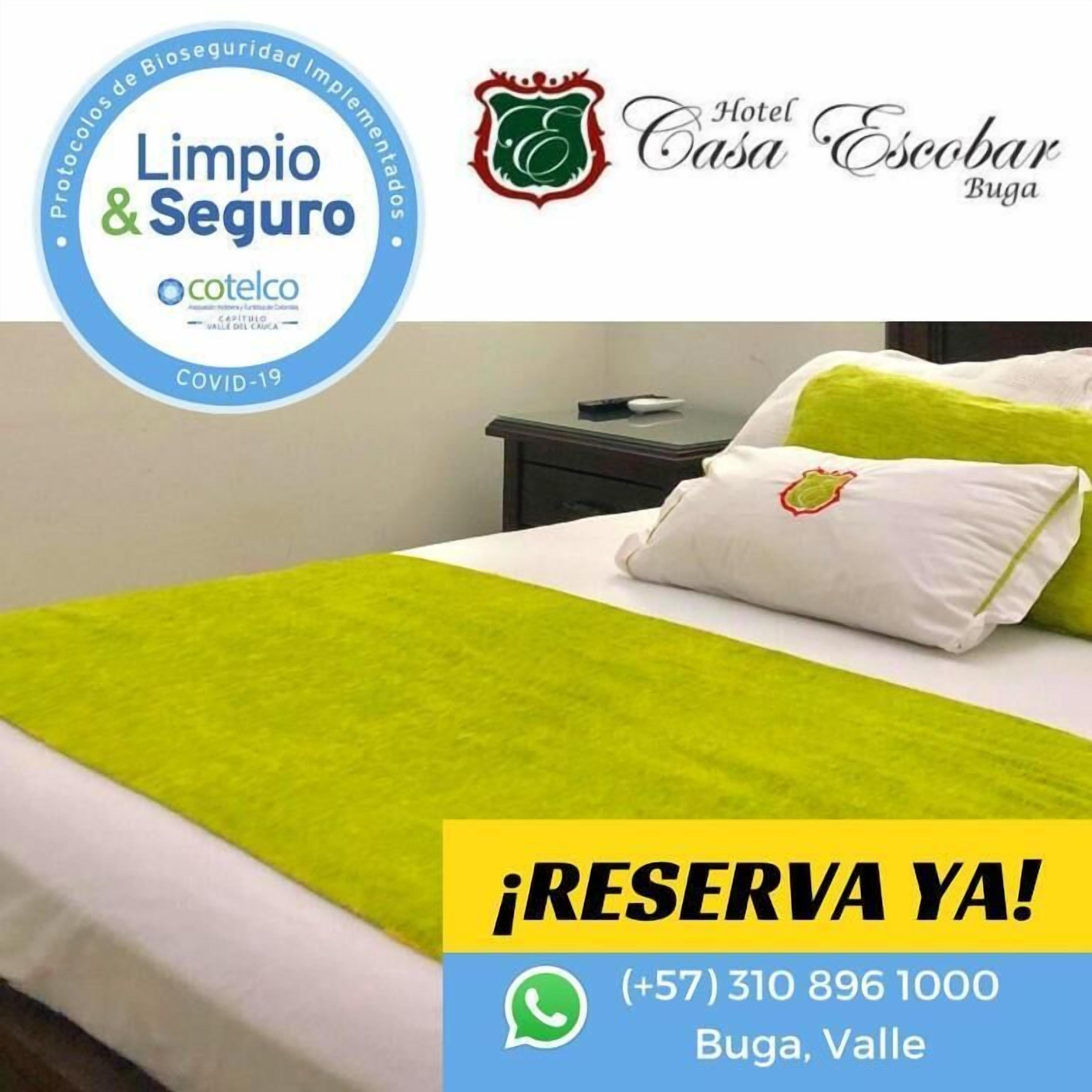 A hotel room with a bed, pillows, and a nightstand. There is a logo for 'Hotel Casa Escobar Buga' and a '¡RESERVA YA!' sign with a phone number.