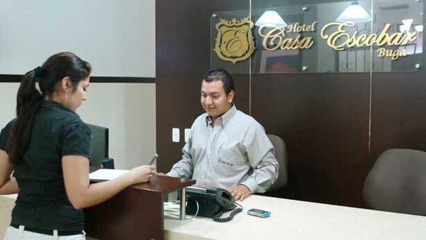 Una mesa de recepción con un hombre sentado detrás, una mujer de pie a la izquierda y un cartel con el nombre del hotel.