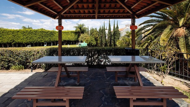 Un área de picnic techada con bancos y mesas, rodeada por un jardín ajardinado con diversas especies de árboles y arbustos.