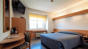 Habitación de hotel con cama, escritorio, silla, televisor, ventana con persianas y aire acondicionado.