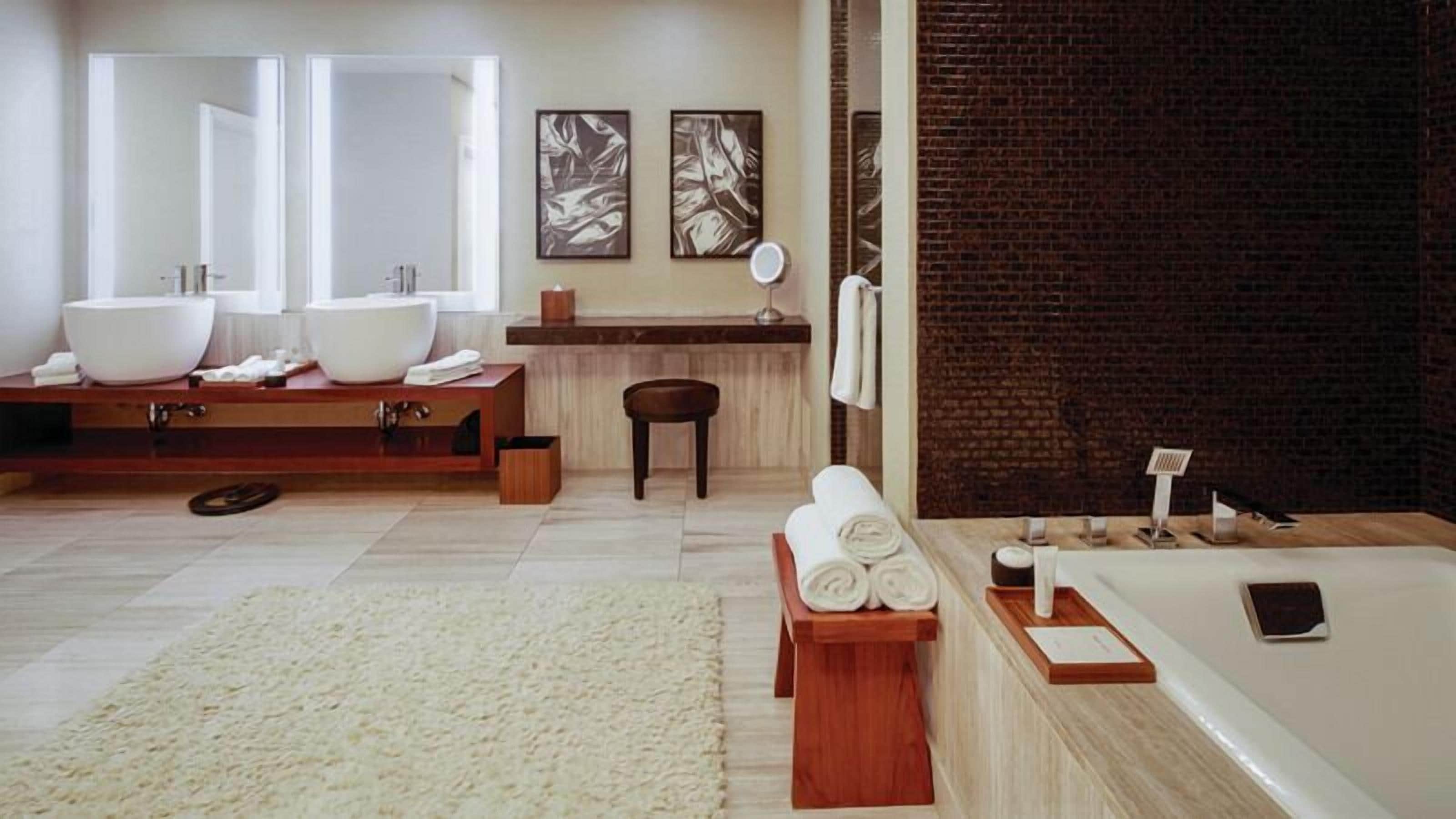 A modern bathroom with two sinks, a large mirror, a wooden vanity, a round wooden stool, a white rug, and a bathtub with a smartphone on the side.