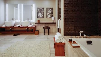 A modern bathroom with two sinks, a large mirror, a wooden vanity, a round wooden stool, a white rug, and a bathtub with a smartphone on the side.