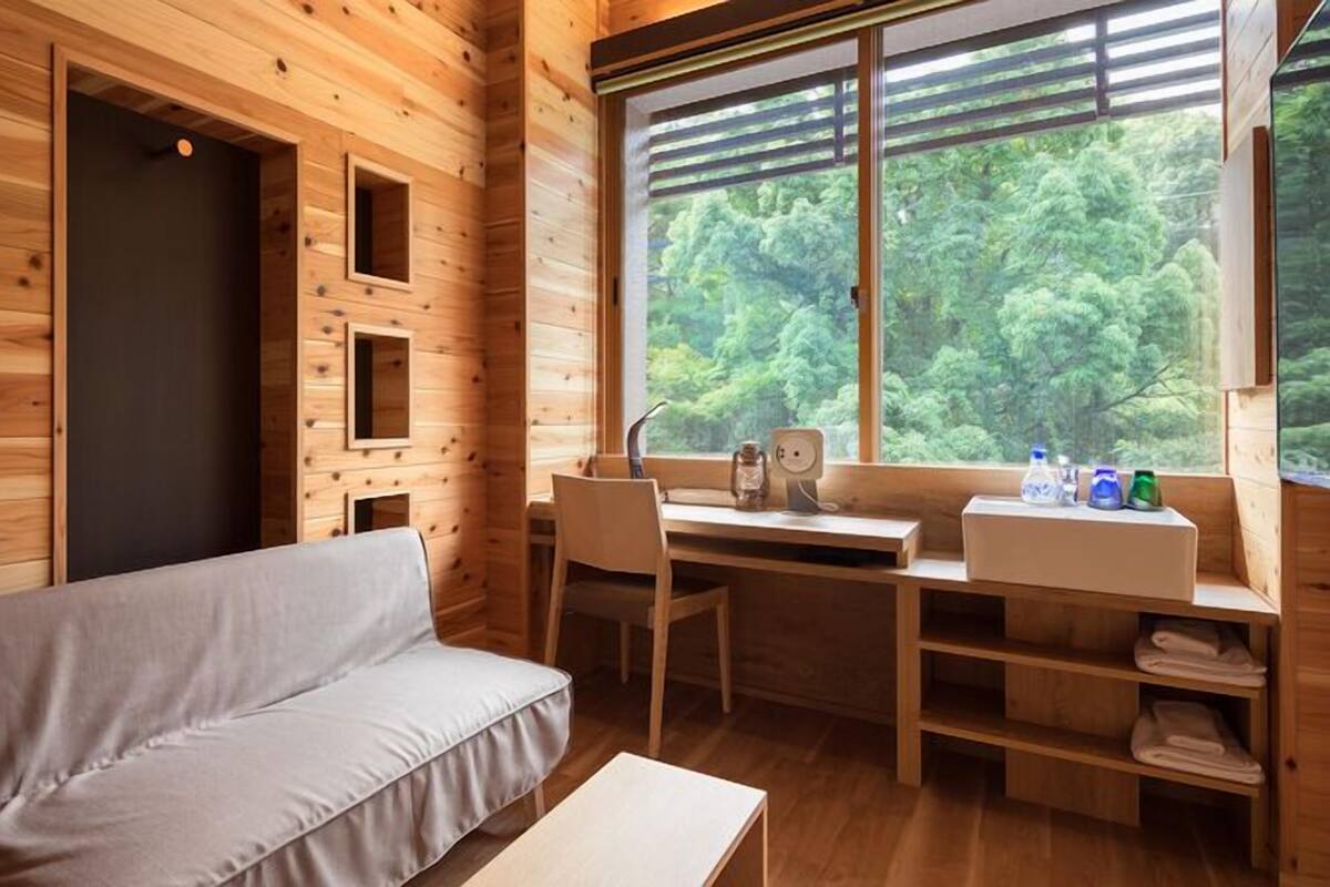 A wooden cabin interior with a sofa, a desk, a window with a view of greenery, and a shelf with towels.