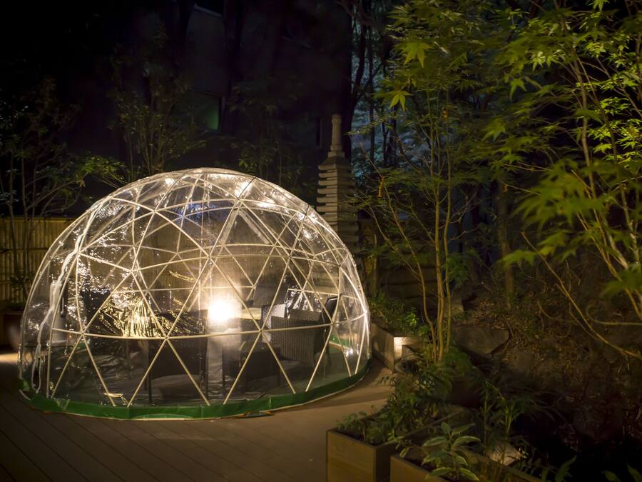 Une structure en forme de dôme géodésique, dotée d’une source de lumière centrale, entourée de verdure et bordée d’un sentier en pierre.