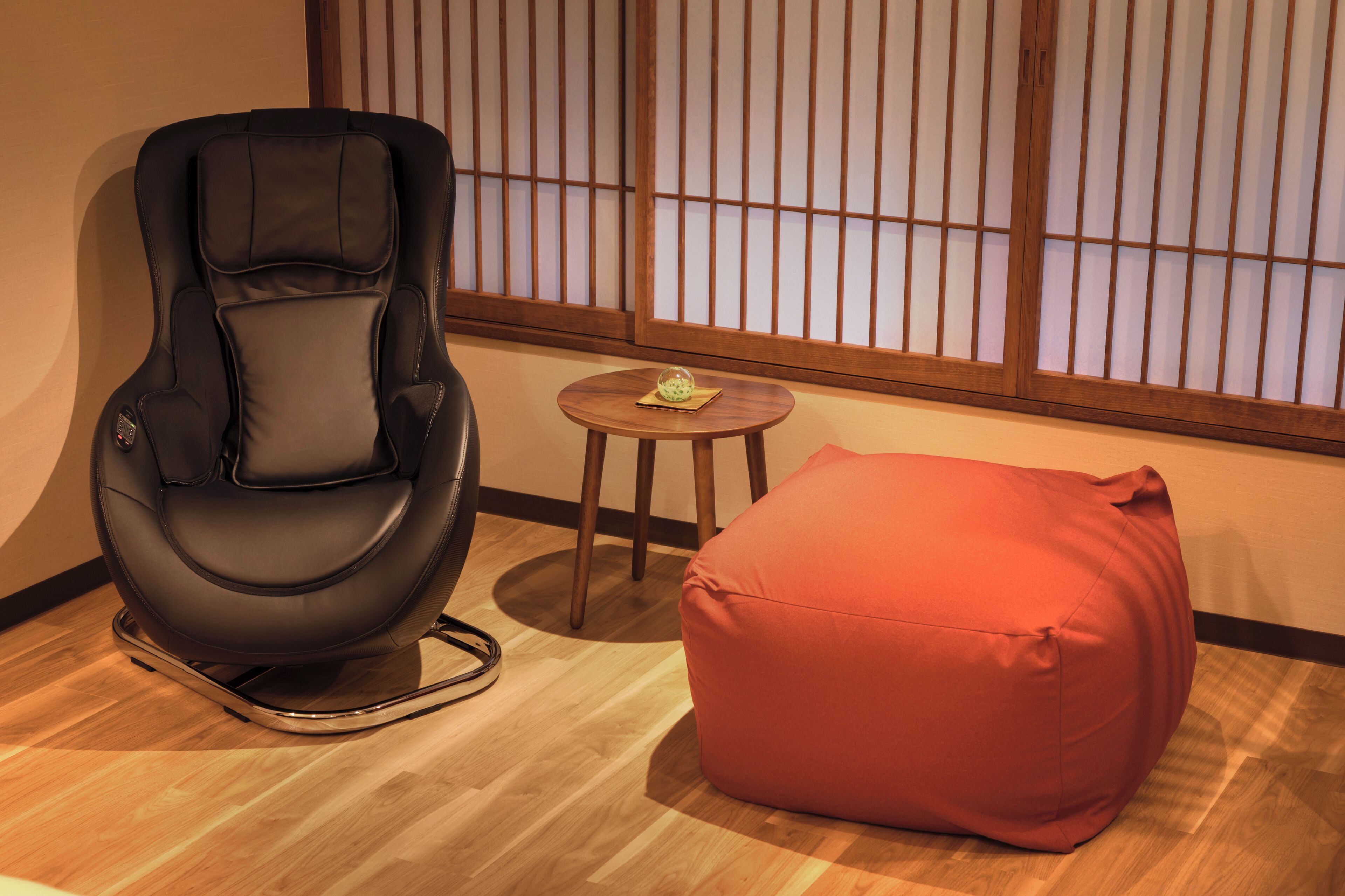 A black massage chair, a small wooden table with a glass object, and a red bean bag chair in a room with wooden flooring and shoji screens.