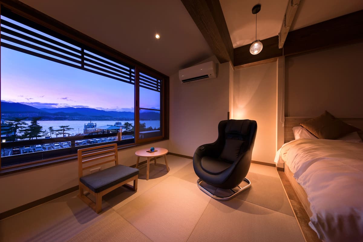 A hotel room with a large window, a bed, a chair, a small table, and an air conditioning unit.