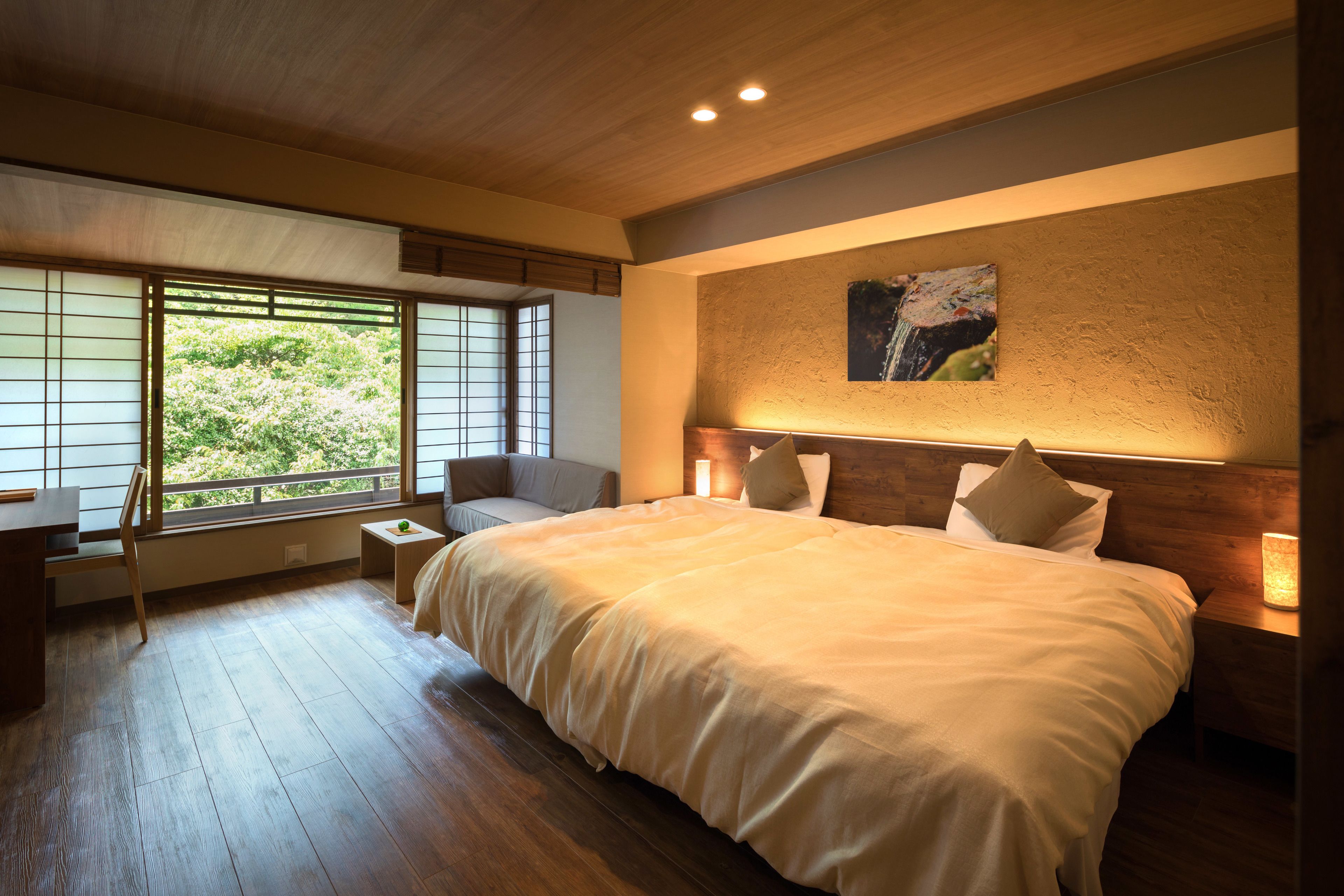 A bedroom with a large bed, a desk, and a chair. There is a window with a view of greenery outside.