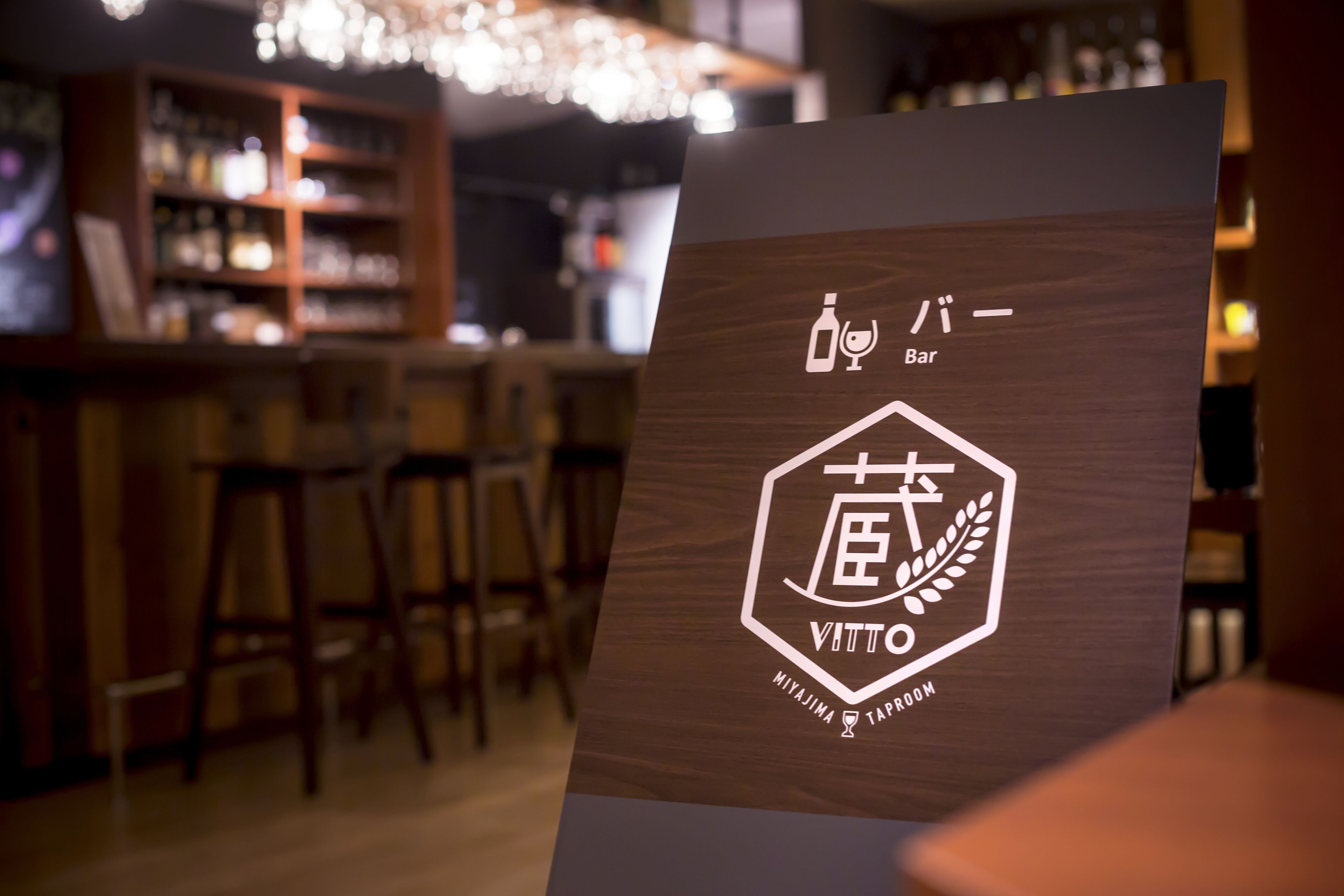 Un letrero de madera con un logotipo y texto que indica una zona de bar, sobre un fondo desenfocado de un bar y asientos.