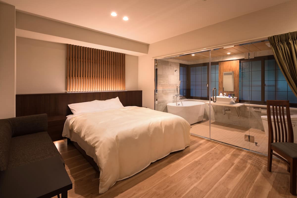 A modern hotel room with a large bed, a sofa, a glass-enclosed bathroom, and a wooden accent wall.
