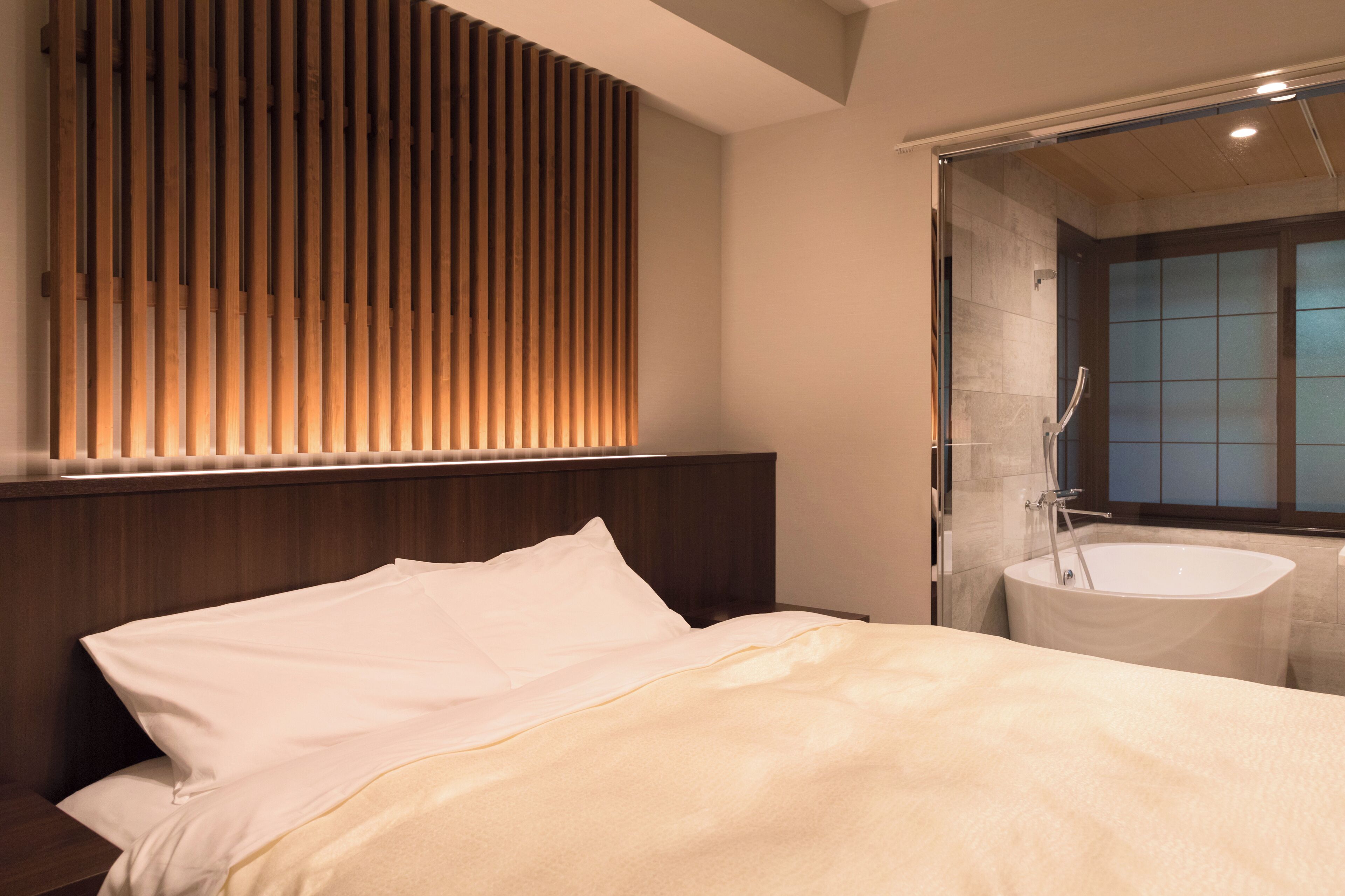 A modern bedroom with a bed, a wooden slat wall, and a bathroom with a freestanding bathtub.