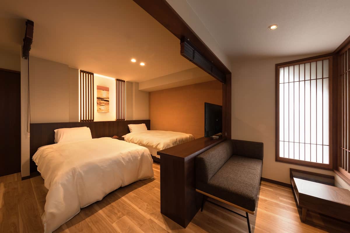 A hotel room with two beds, a sofa, a TV, and a large window with vertical blinds.
